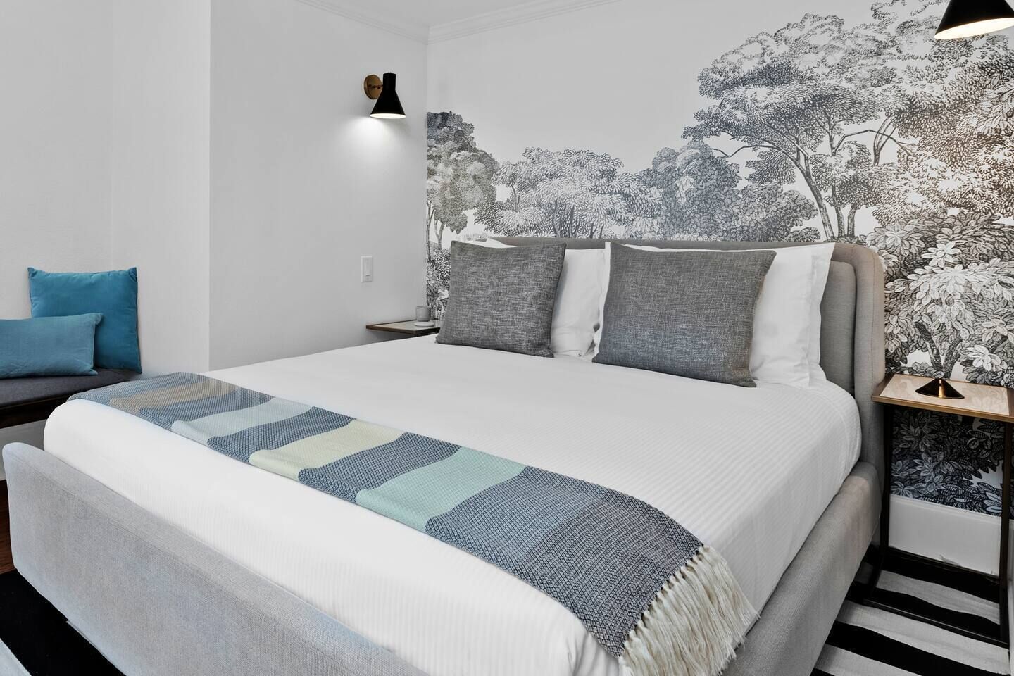 A bed with a patterned blanket, a nightstand with a lamp, and a wall with a nature-themed mural.