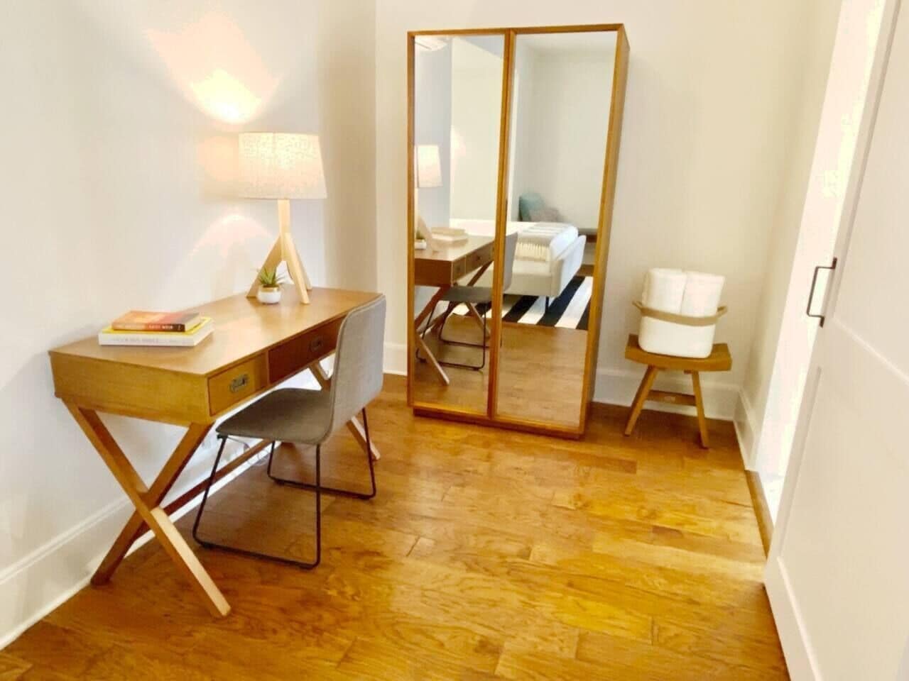 A room with a wooden desk, a chair, a lamp, a mirror, and a small stool.