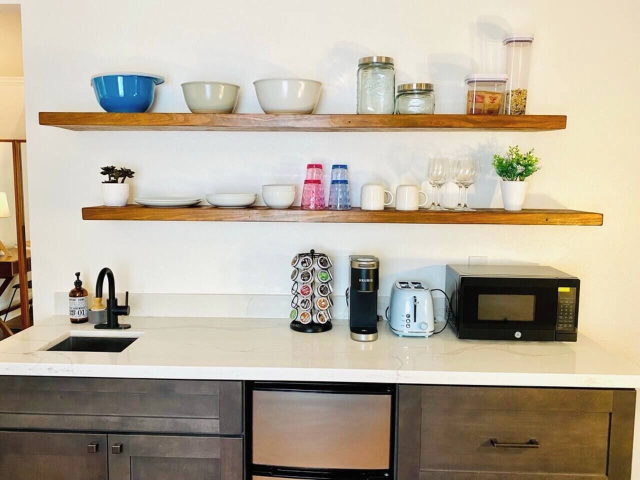 A kitchen with a white countertop, a sink, a coffee maker, a microwave, and shelves above holding bowls, jars, and a potted plant.