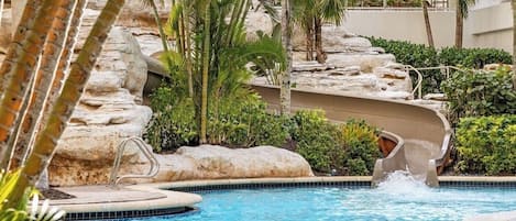 Uma piscina com escorrega aquático, rodeada por palmeiras e pedras.