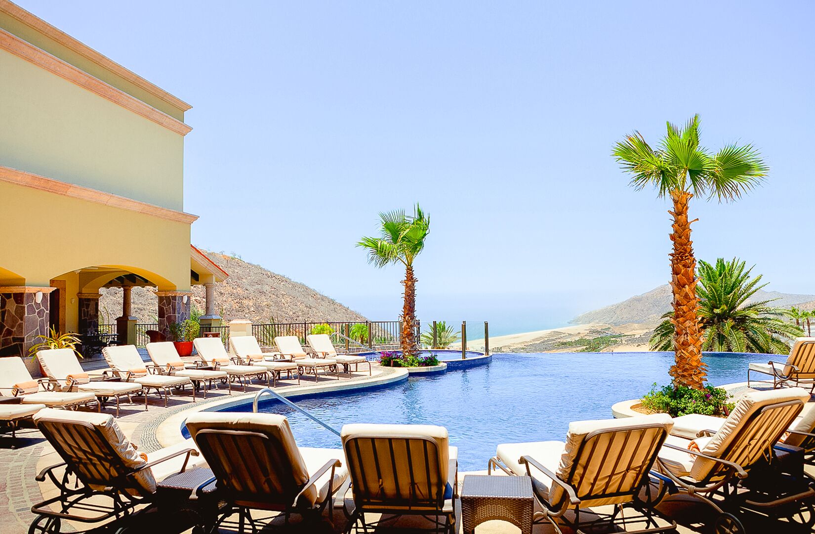 A poolside area with lounge chairs and palm trees, overlooking a beach and mountains.