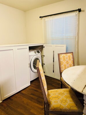 A compact kitchen with a washing machine, a dining table with a chair, and a window with blinds. - Pet-Friendly Home Near Dragon Con & Downtown (Atlanta)