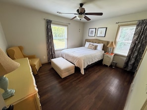 Quarto com cama, mesa de cabeceira, cadeira, janela com cortinas e ventilador de teto.