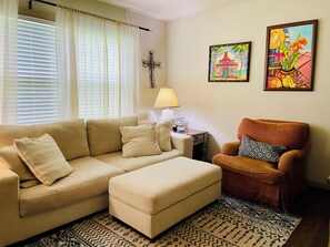 Uma sala de estar com um sofá bege, um banquinho, um tapete estampado, uma mesa lateral com abajur e duas obras de arte emolduradas na parede.