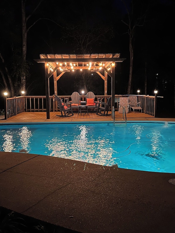 Una piscina con muelle de madera y sillones de descanso, rodeada por una pérgola de madera con luces colgantes.