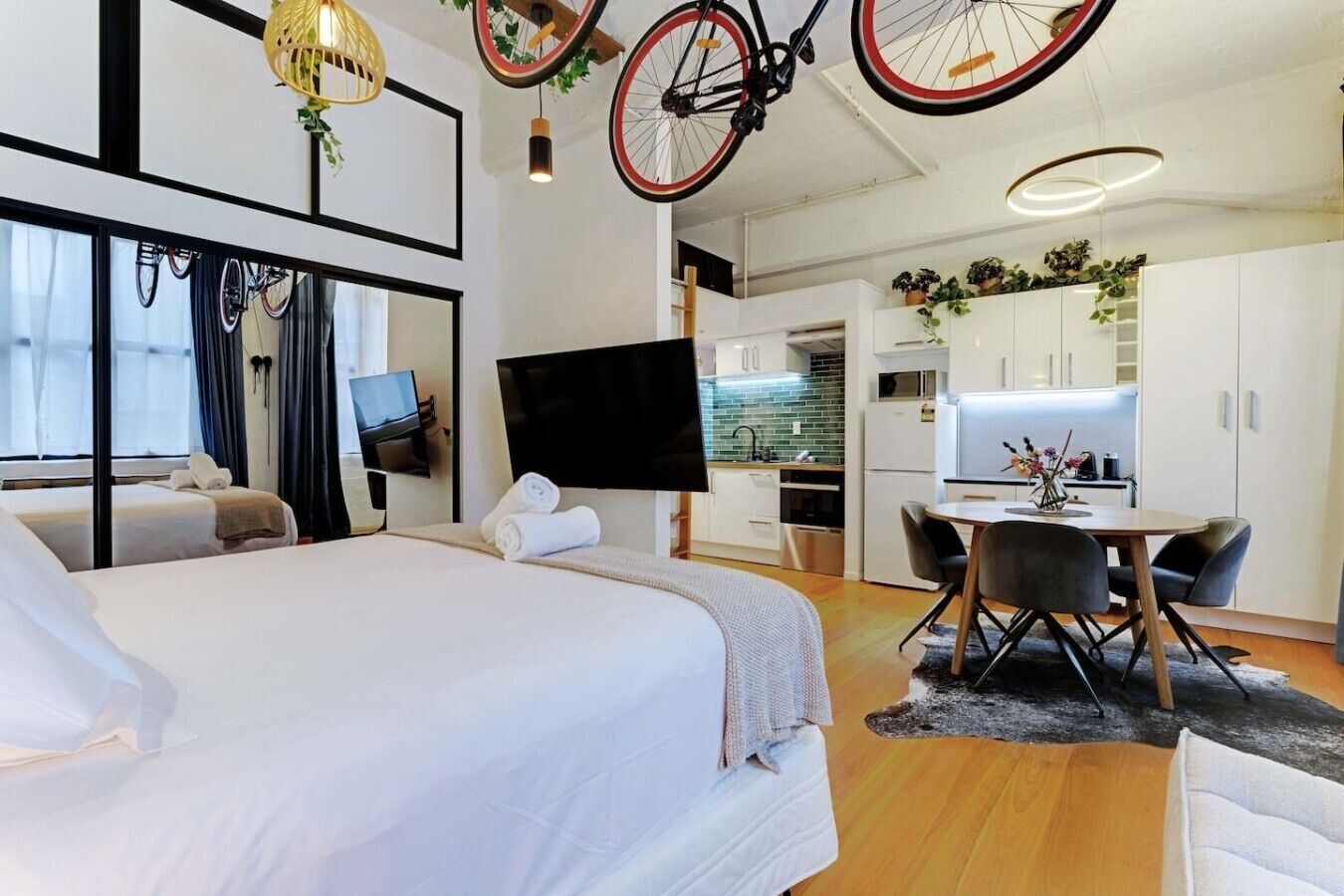 Een moderne hotelkamer met een groot bed, een zithoek met een tafel en stoelen, een keuken met witte kasten en een televisie aan de muur.