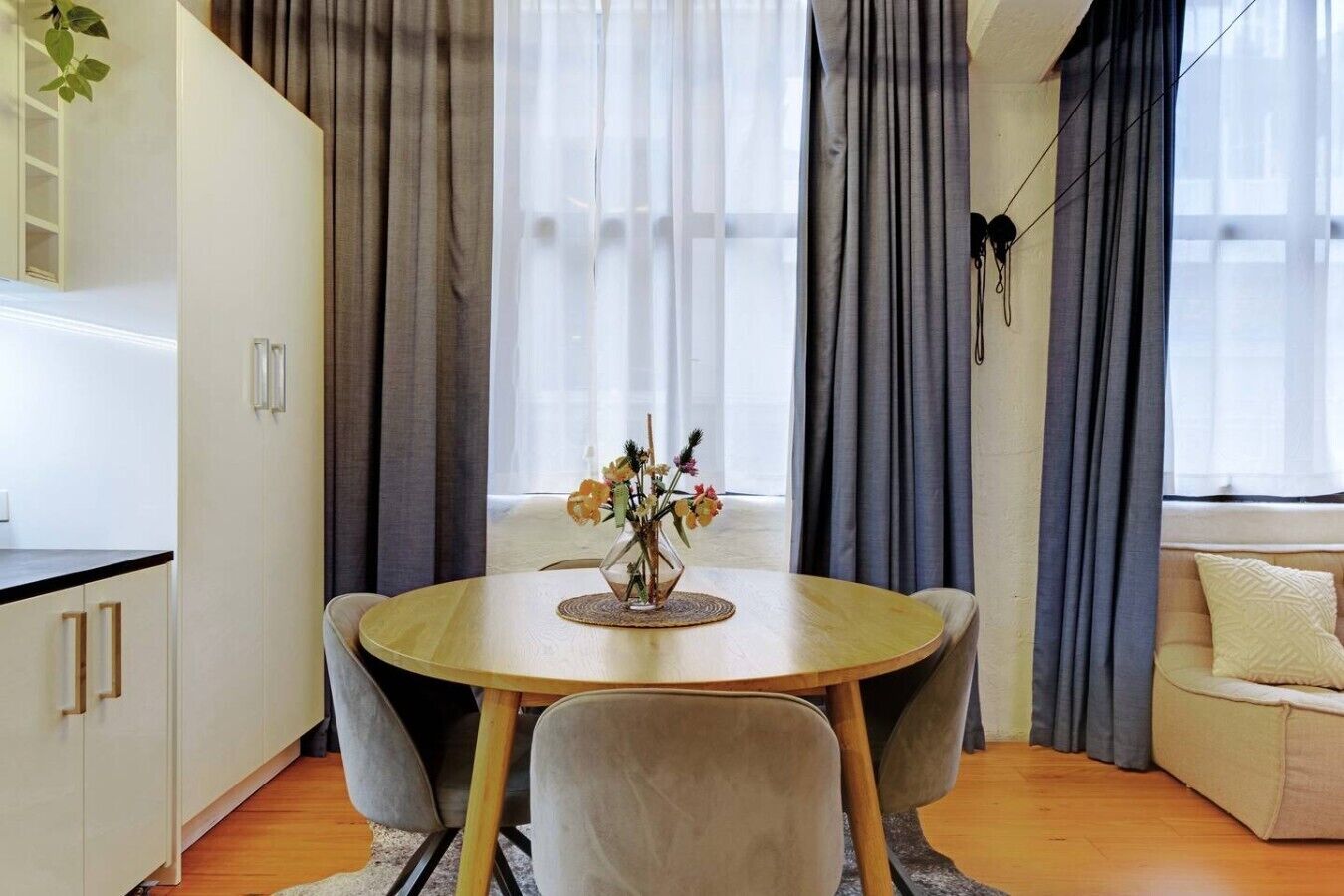 A modern living room with a round wooden dining table, grey upholstered chairs, a beige sofa, and a window with grey curtains.