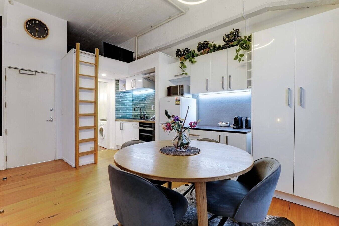 A compact kitchen with white cabinets, a wooden dining table with chairs, and a small plant on the counter.