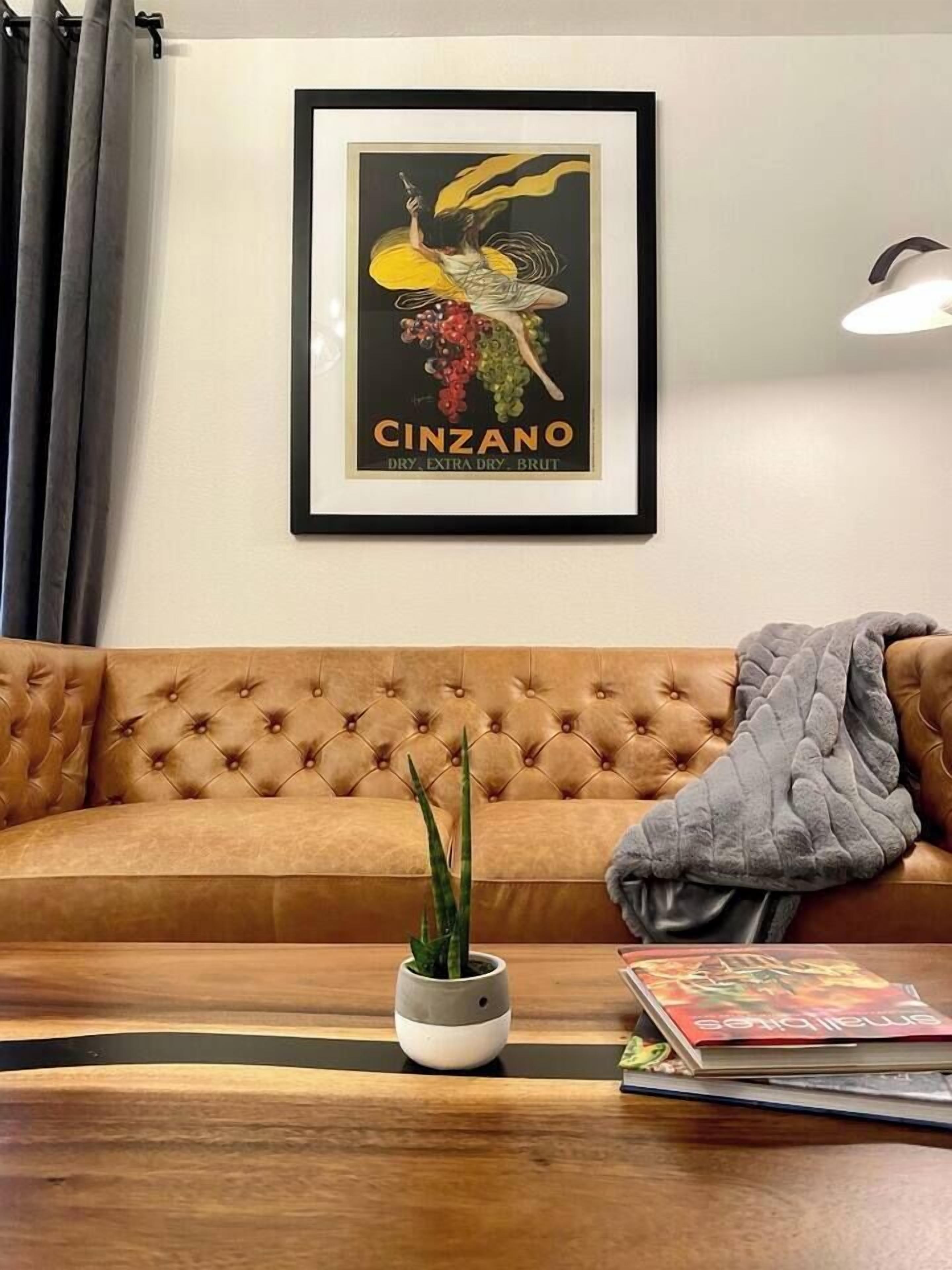A framed poster of a woman holding a bottle, a brown tufted sofa, a small potted plant, and a magazine on a wooden coffee table.