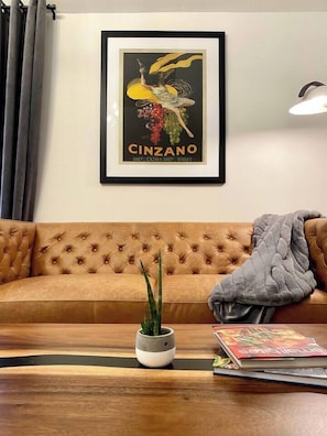A framed poster of a woman holding a bottle, a brown tufted sofa, a small potted plant, and a magazine on a wooden coffee table.