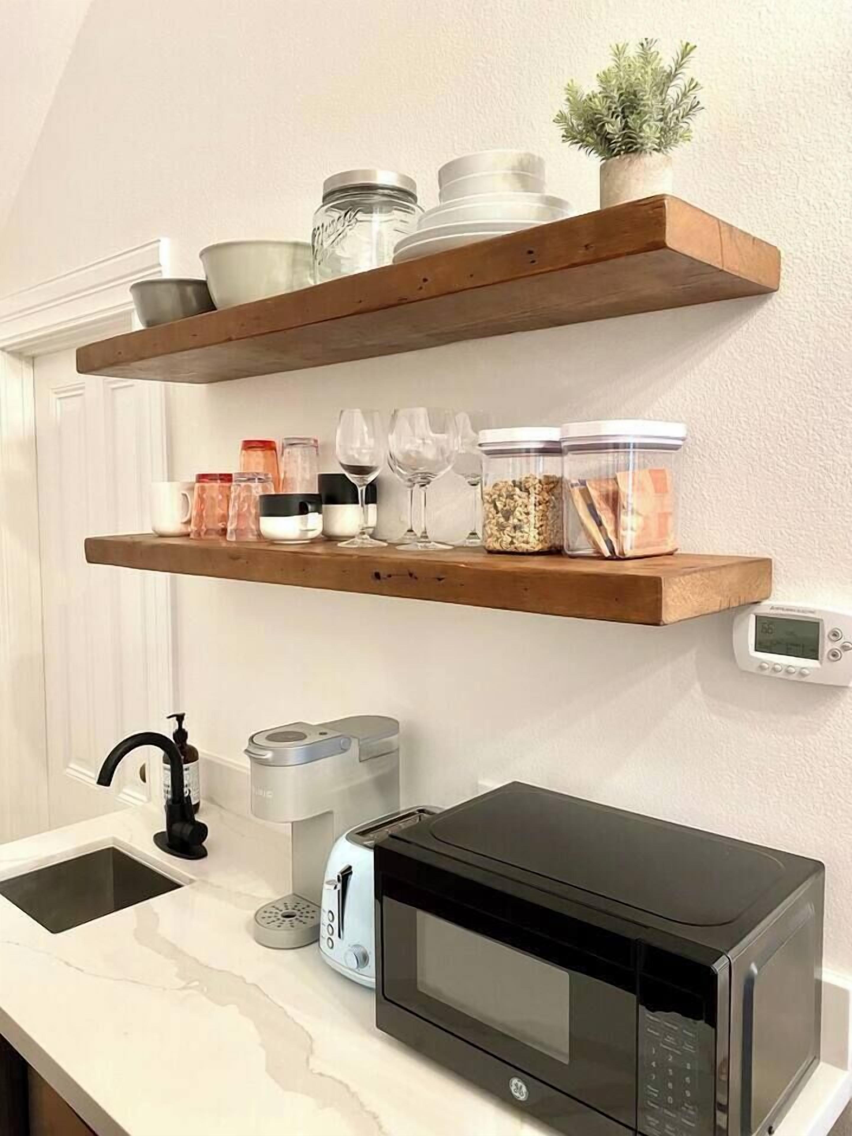 A kitchen with a microwave, toaster, and a wooden shelf above the counter holding jars and glasses.