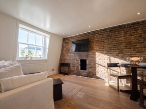 A modern living room with a brick fireplace, a flat-screen TV mounted on the wall, a dining area with a table and chairs, and a large window providing natural light. - South Ken Rooftop Loft, Treetop Views, Hyde Park (London)