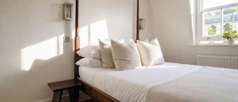A bedroom with a wooden bed, white bedding, a nightstand, a wall-mounted light, and a window with a view.