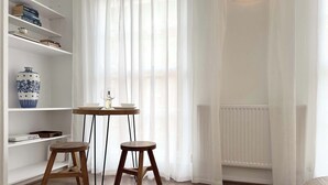 Une pièce avec une bibliothèque blanche, une petite table en bois accompagnée de deux tabourets, un vase et un radiateur.