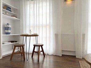 Een kamer met een witte boekenkast, een klein houten tafeltje met twee krukken, een vaas en een radiator.