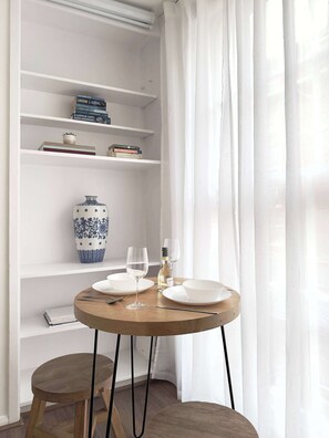 A dining area with a round wooden table, white plates, wine glasses, and a blue and white vase on a shelf. - Chic Marble Arch w/ 100" Cinema & Premium Mattress (London)