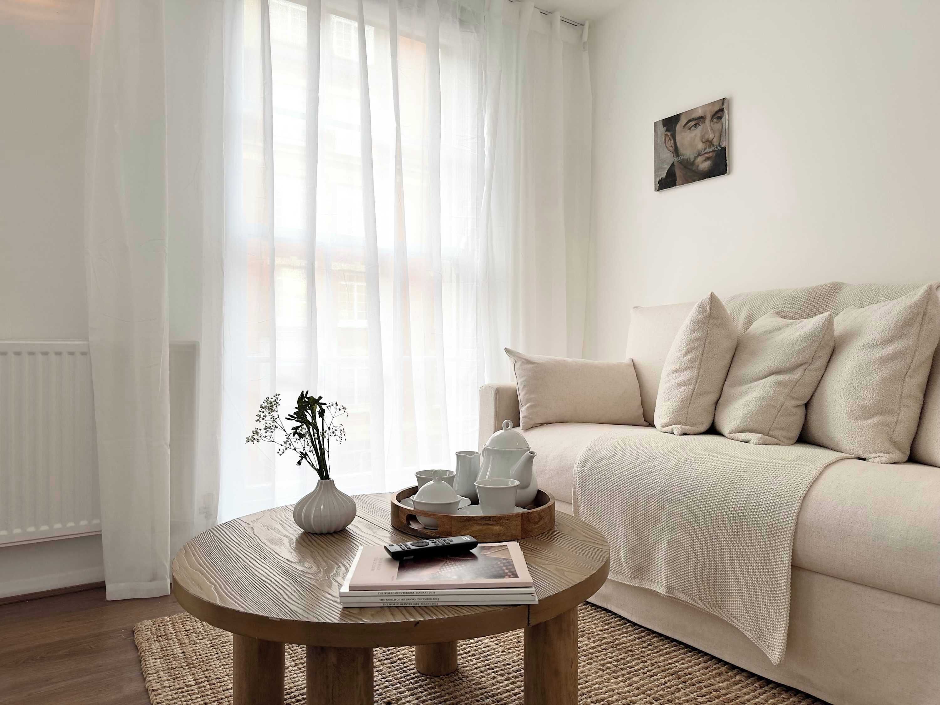 Ein Wohnzimmer mit einem runden Holztisch, einer Couch, einer weißen Vase mit Blumen und einem Bild an der Wand.