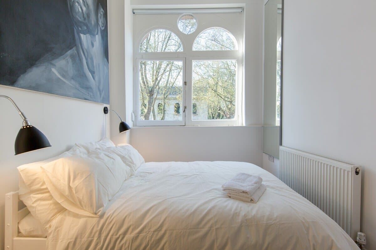A bedroom with a bed, a window with a view, a painting on the wall, and a radiator.
