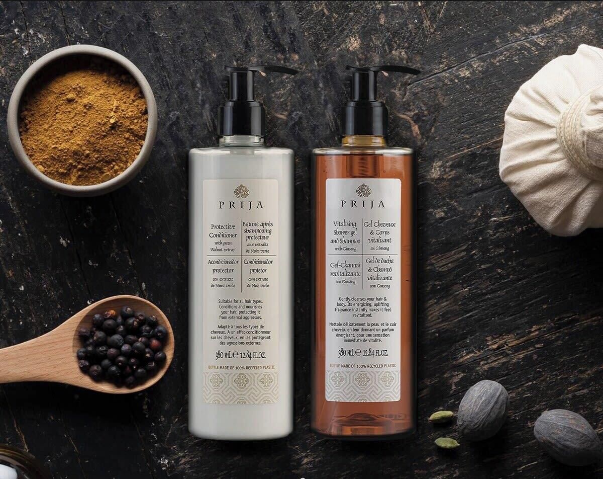 Deux bouteilles de produits PRIJA, un bol de poudre brune, une cuillère en bois décorée de baies et quelques amandes sur une surface en bois sombre.