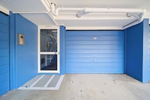 A blue garage door with a metal door and a small window, a white pipe on the ceiling, and a concrete ground. - Passage Panorama - 3BD Seaview Retreat (Caloundra Region)