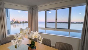Quarto de hotel com uma mesa de jantar, cadeiras e vista para a água e o horizonte urbano.