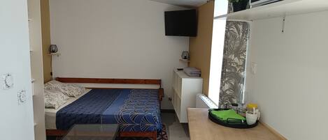 A compact room with a bed, a small table, a TV mounted on the wall, and a window with curtains.