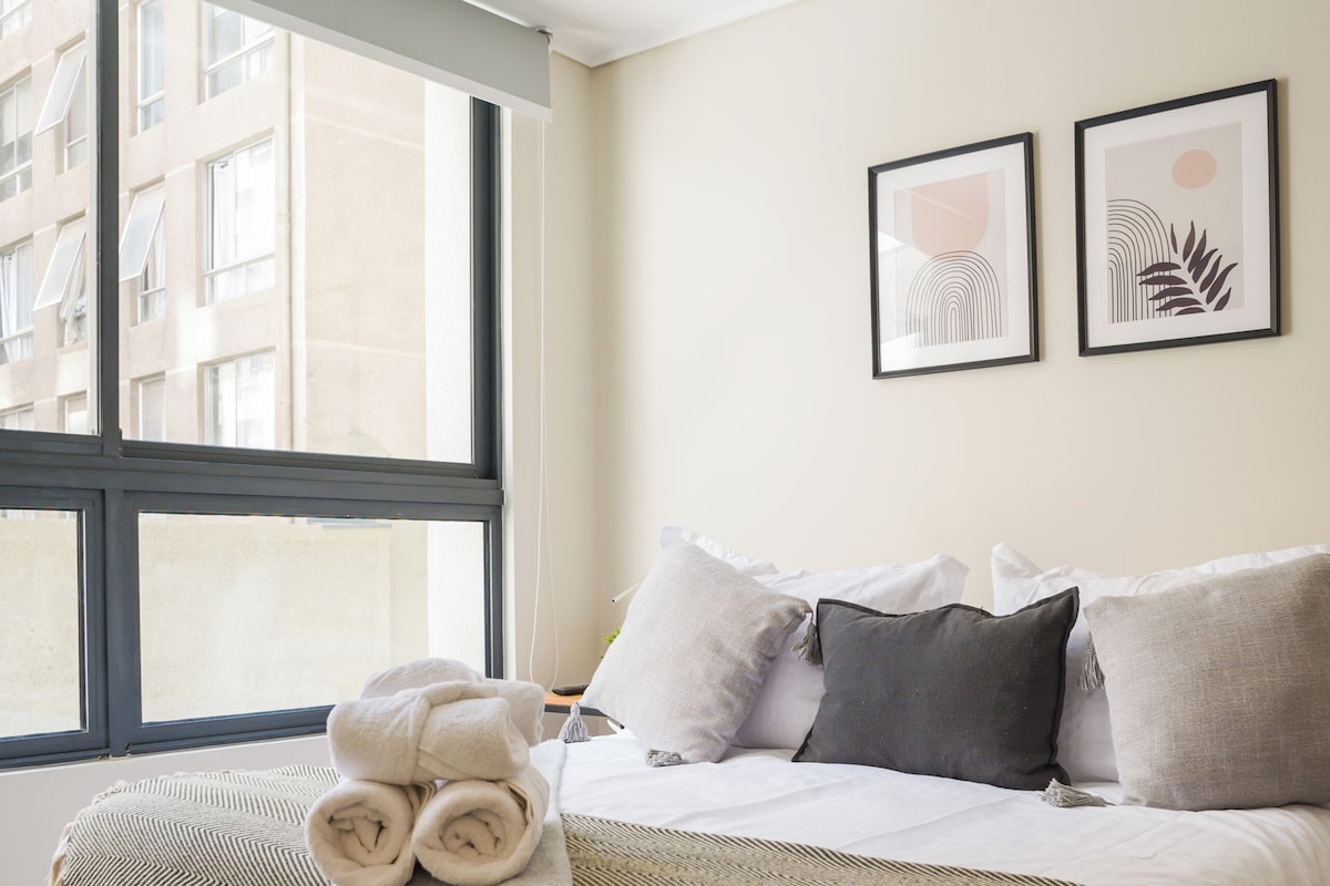 Ein Hotelzimmer mit einem großen Fenster, einem Bett mit vielen Kissen, einer Decke und zwei gerahmten Kunstwerken an der Wand.