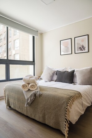 Ein Hotelzimmer mit einem großen Fenster, einem Bett mit vielen Kissen, einer Decke und zwei gerahmten Kunstwerken an der Wand.