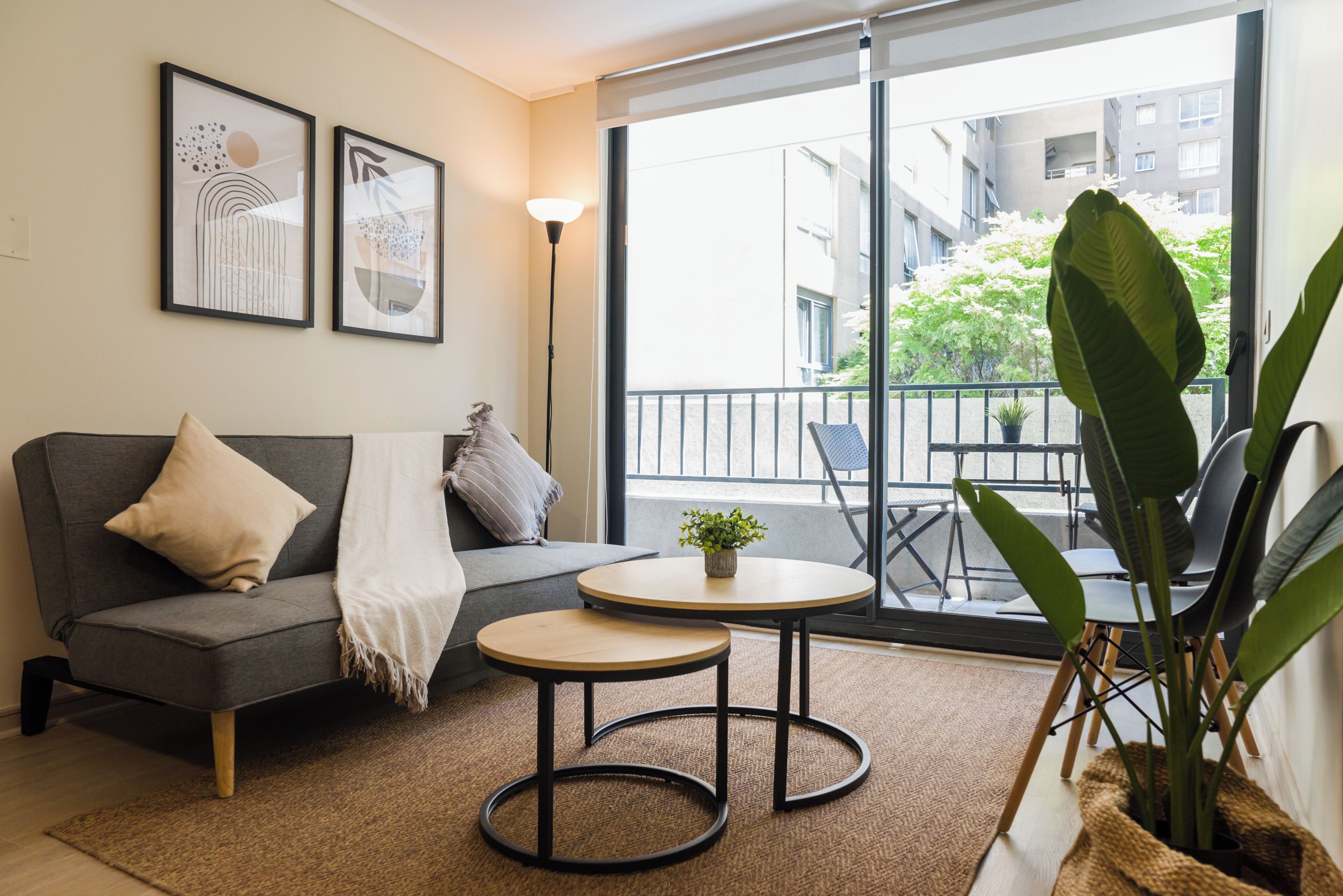 A modern living room with a grey sofa, two small tables, a potted plant, and a balcony with outdoor furniture.
