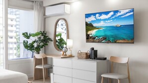 Ein modernes Hotelzimmer mit Bett, Schreibtisch mit Stuhl, großem Fenster mit Vorhängen, Zimmerpflanze, Globus, Lampe, gewebtem Korb und einem gerahmten Bild von einem Strand.