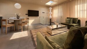 Ein modernes Wohnzimmer mit einer Couch, einem Couchtisch und einem Essbereich mit Stühlen und Tisch. Ein Flachbildfernseher ist an der Wand montiert und über der Couch befindet sich ein Oberlicht.