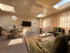 Un salon moderne avec un canapé, une table basse et un coin repas comprenant des chaises et une table. Un téléviseur à écran plat est fixé au mur, et une lucarne se trouve au-dessus du canapé.