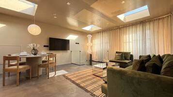 A modern living room with a dining area, a flat-screen TV, and a large window with curtains.