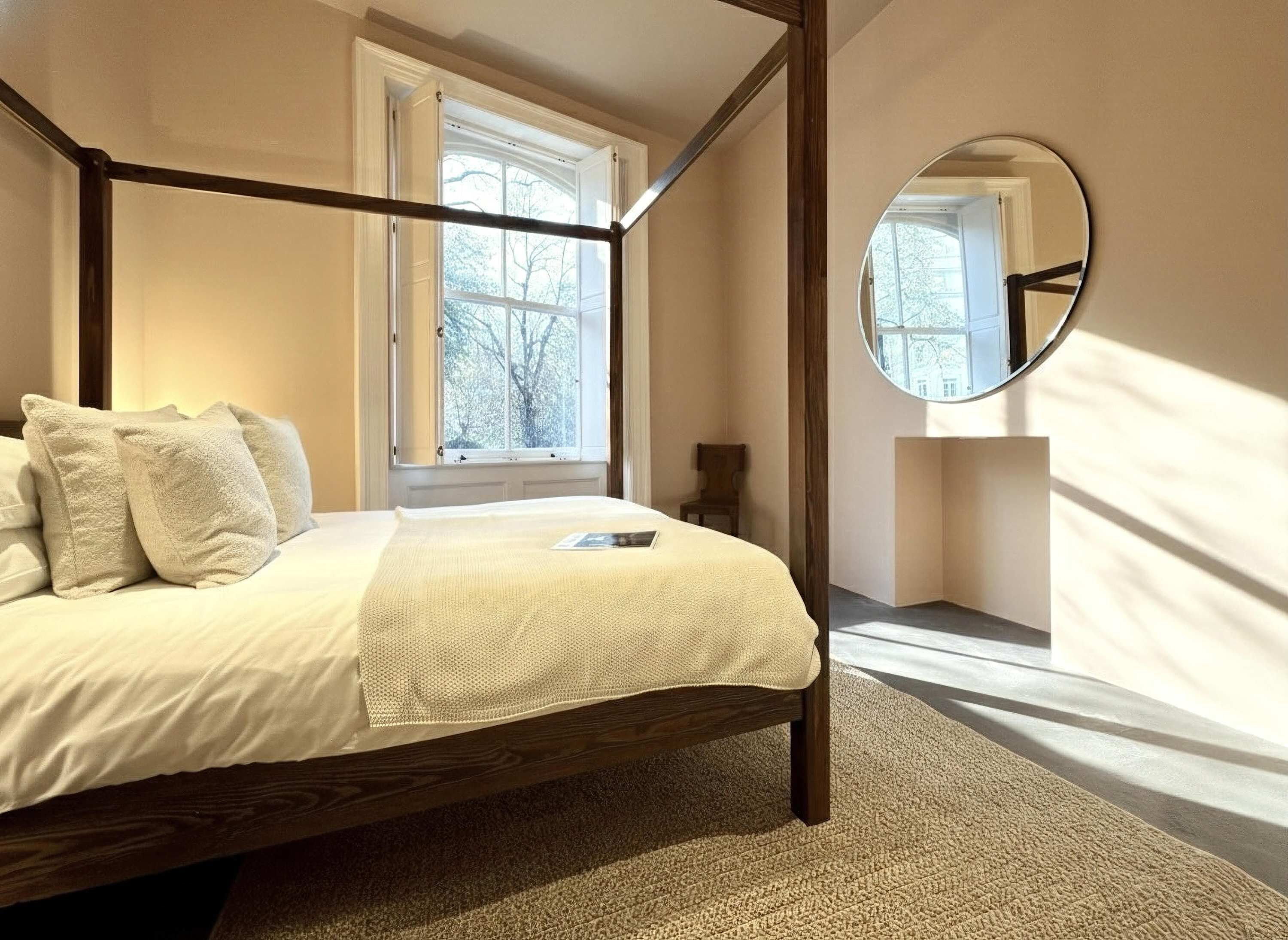 A four-poster bed with a white bedspread and pillows, a round mirror on the wall, and a window with a view of trees.