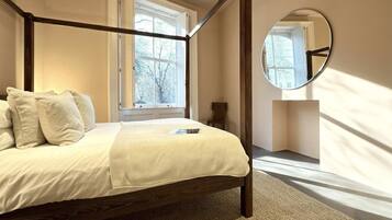 A four-poster bed with a white bedspread and pillows, a round mirror on the wall, and a window with a view of trees.