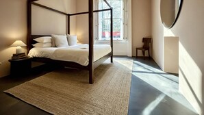 Ein Bett mit Baldachin und Holzrahmen, ein Nachttisch mit Lampe, ein Sessel und ein großer Spiegel an der Wand.