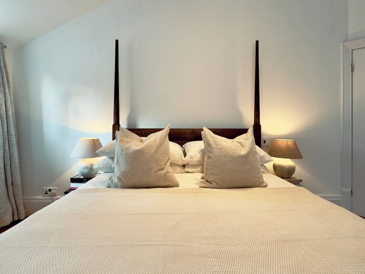 A neatly made bed with a white duvet and two pillows, a bedside lamp, and a nightstand with books and a lamp.