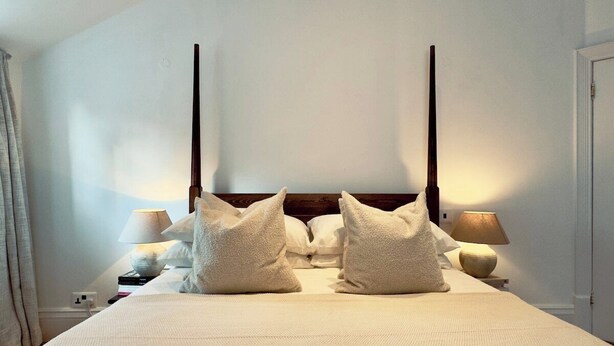 A neatly made bed with a white duvet and two pillows, a bedside lamp, and a nightstand with books and a lamp. - Grand 2BR Penthouse | 100" Cinema | Hyde Park (London)