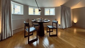 A dining area with a round table, four chairs, and a pendant light. There are three windows with curtains, a radiator, and a wooden floor. - Grand 2BR Penthouse | 100" Cinema | Hyde Park (London)