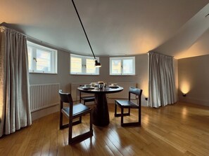 Ein Essbereich mit einem runden Tisch, vier Stühlen und einer Pendelleuchte. Drei Fenster mit Vorhängen, ein Heizkörper und ein Holzboden sind ebenfalls vorhanden.