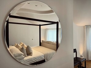 A circular mirror reflecting a four-poster bed with pillows, a bedside table, and a window with curtains. - Grand 2BR Penthouse | 100" Cinema | Hyde Park (London)