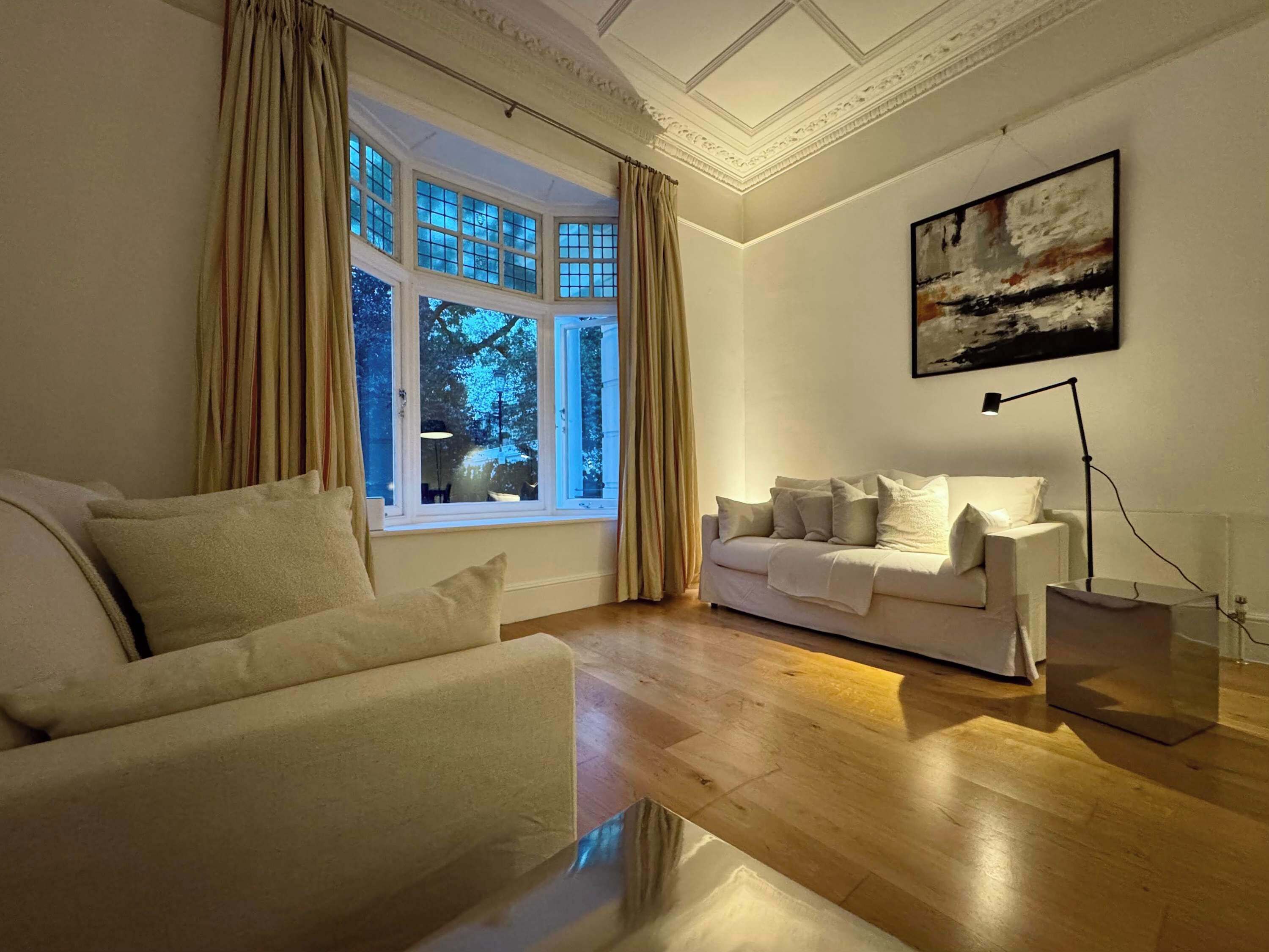 A living room with a sofa, a white armchair, a large window with curtains, a painting on the wall, and a floor lamp.