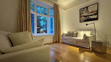 A living room with a sofa, a white armchair, a large window with curtains, a painting on the wall, and a floor lamp.