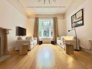 A spacious room with two white sofas, a fireplace, a large window with curtains, a floor lamp, and a painting on the wall. - Grand South Ken Sanctuary | Fire Place | Courtyard (London)