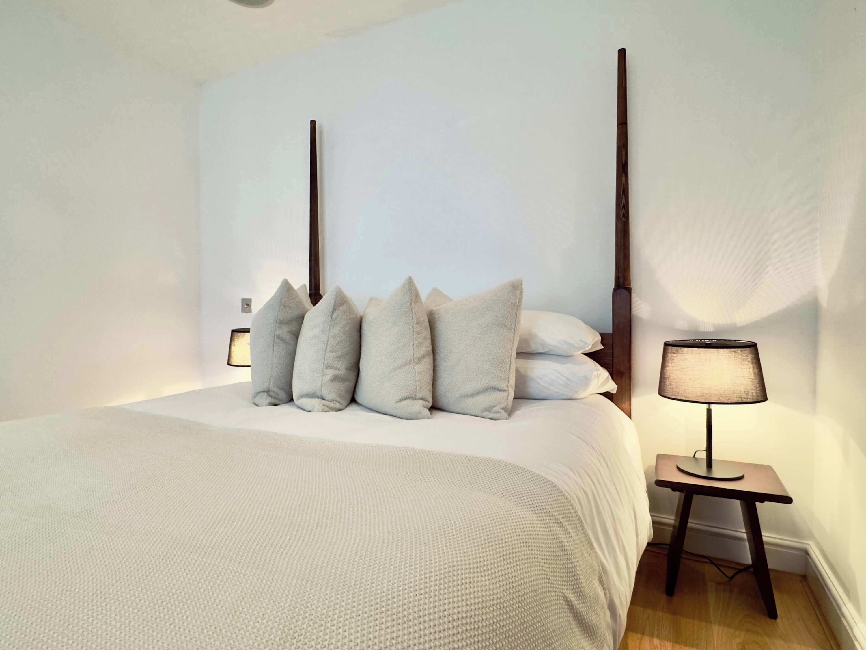 A bedroom with a bed, pillows, a bedside table with a lamp, and a wooden floor.