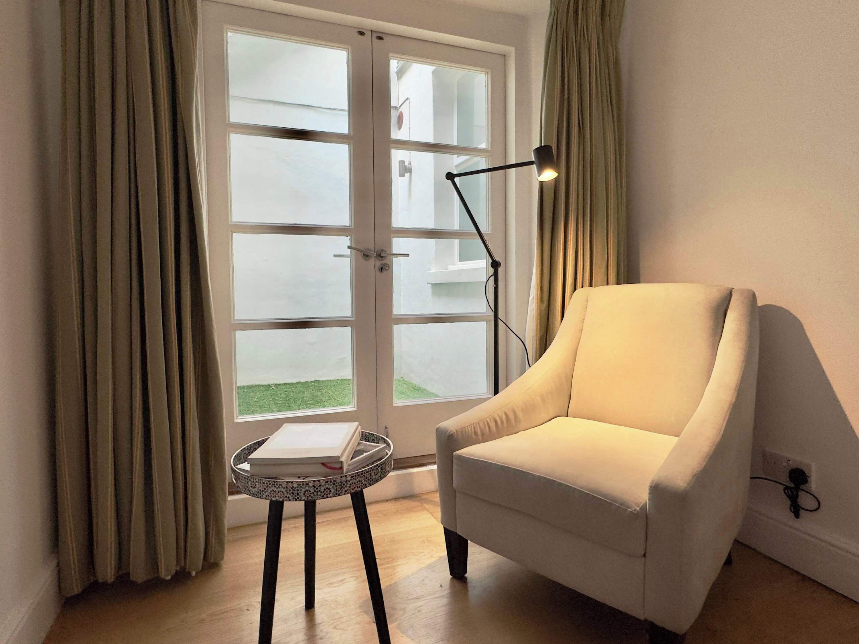 A room with a white armchair, a small round table with a book, a floor lamp, and a glass door leading outside.