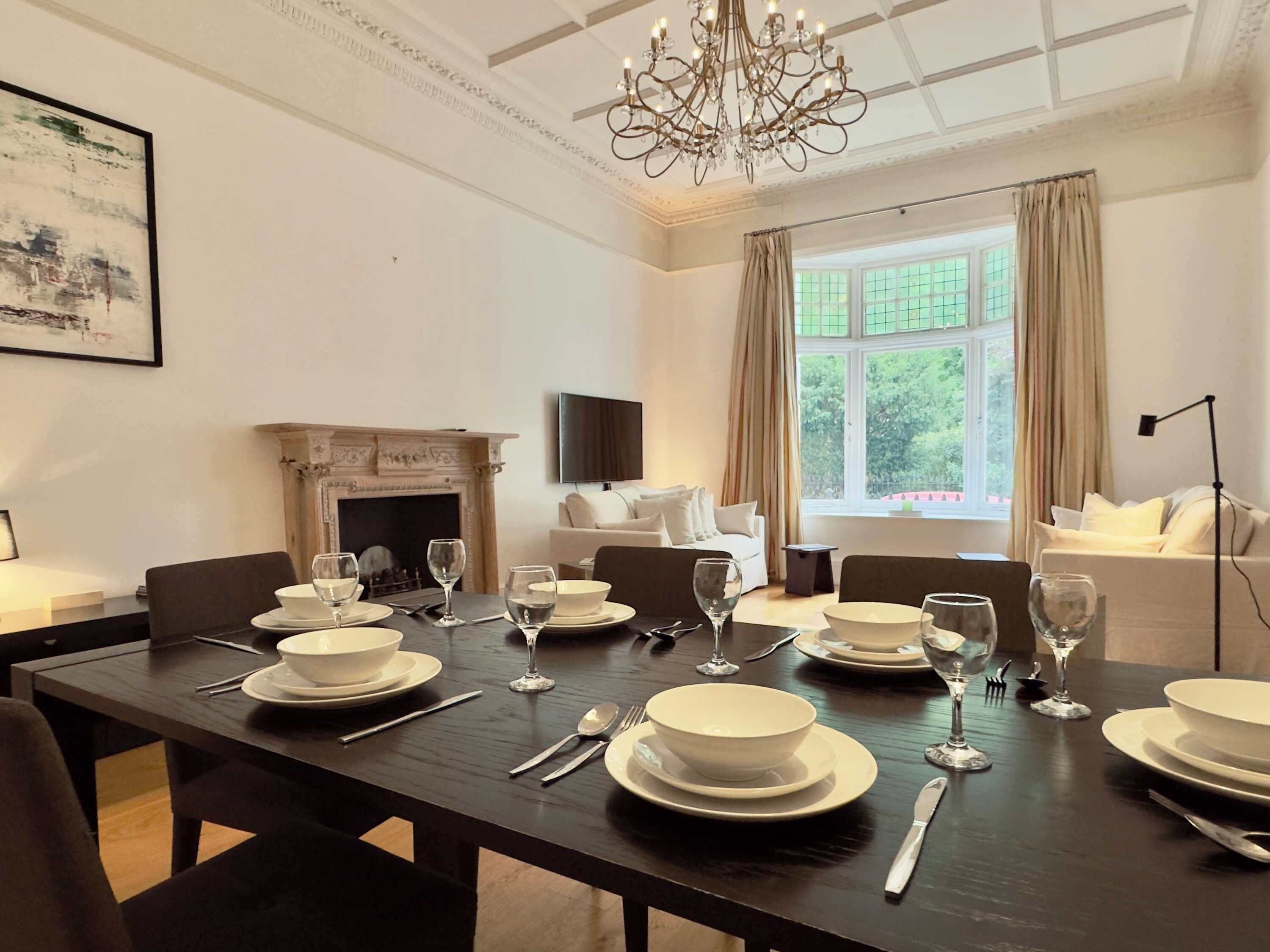 A dining room with a long table set for a meal, a fireplace, a sofa, and a large window with curtains.