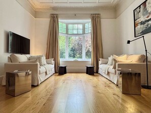 Un salon avec deux canapés, un téléviseur, une lampe sur pied et une grande fenêtre avec des rideaux.