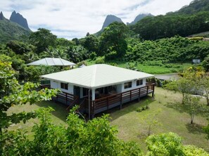 A single-story house with a flat roof, surrounded by lush greenery and mountains in the background.