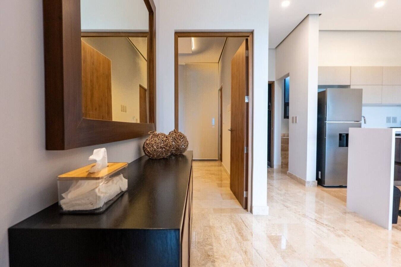 Un moderno vestíbulo de hotel con piso de mármol, recepción de madera, un espejo grande y vista al área de cocina.